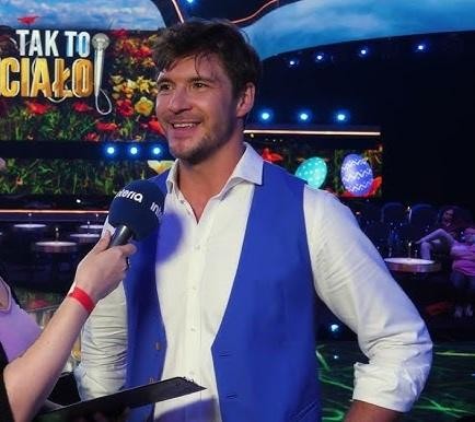 Alt: Mikołaj Roznerski na scenie teatralnej, w trakcie występu, z intensywnym wyrazem twarzy.