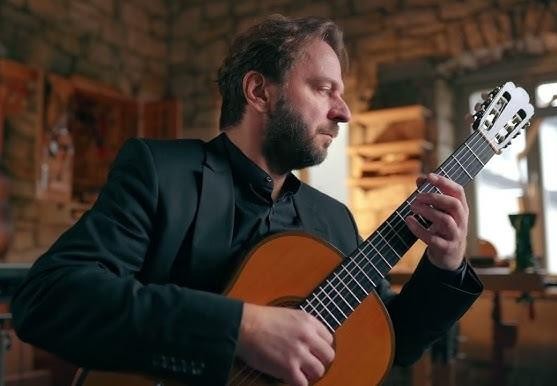 Marcin Dylla podczas występu na scenie, z intensywnym skupieniem i dynamicznym ujęciem dłoni na gitarze, oświetlony reflektorami.