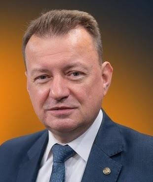 Mariusz Błaszczak jako minister obrony narodowej, w kontekście militarnym, np. podczas inspekcji wojsk, w pobliżu pojazdów wojskowych (czołg) lub na wydarzeniu związanym z przemysłem obronnym, ukazany w sposób dynamiczny i autorytatywny.